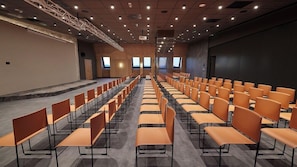 Un salón de conferencias con filas de sillas, un escenario y una pantalla grande.