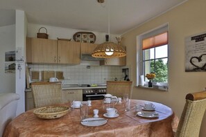 Uma área de refeições com mesa redonda posta para quatro pessoas, cercada por cadeiras de vime, um lustre e uma janela com vista para o verde.