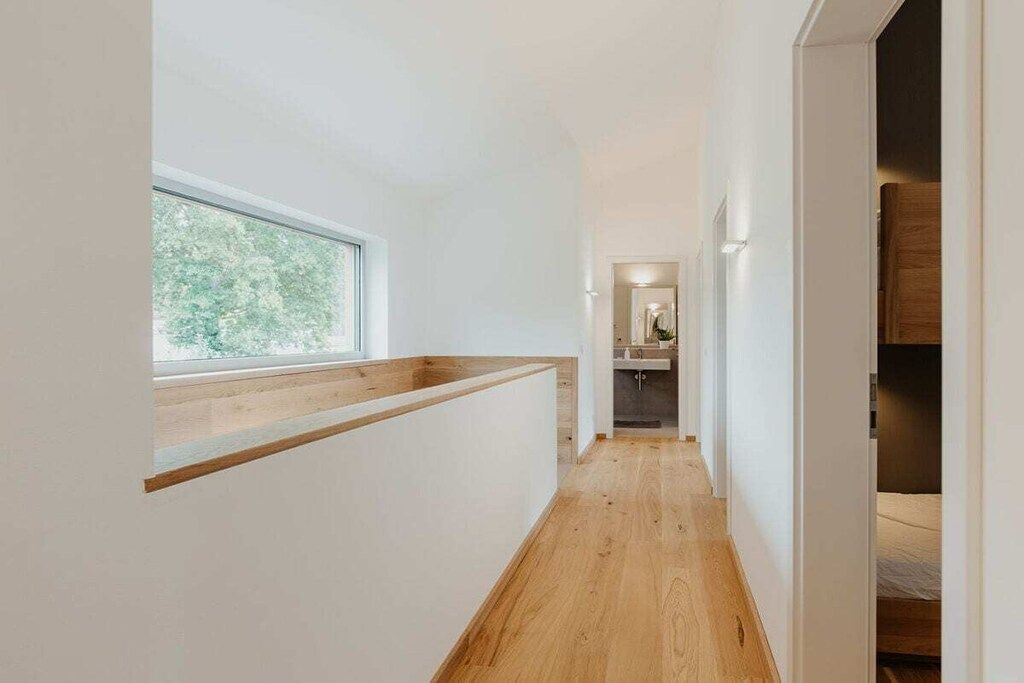 A narrow hallway with a wooden floor, a window on the left, and a bed visible in the distance.