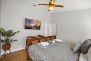 Quarto com cama, mesa de cabeceira, televisão na parede, ventilador de teto, porta e planta em vaso.