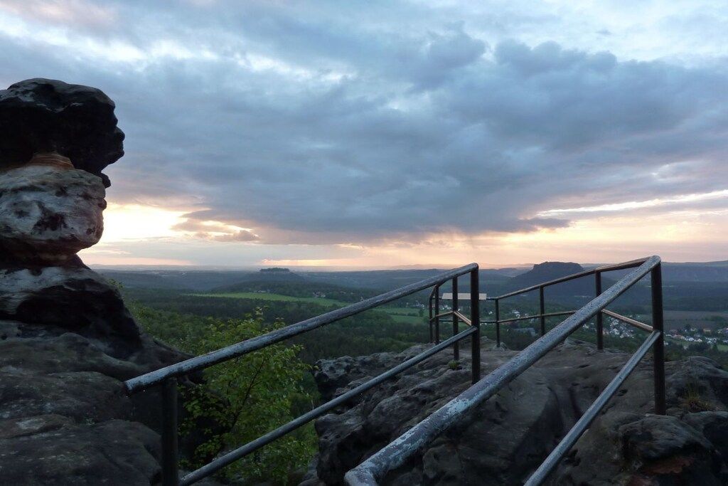 A metal staircase leading up a rocky hill with a metal railing, overlooking a valley with distant hills and a cloudy sky.