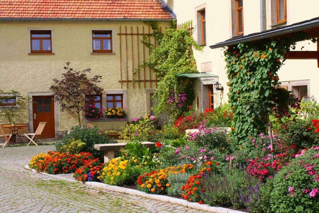 Een straat met cobblestones die leidt naar een gebouw met een rood pannendak, omgeven door een tuin met verschillende bloeiende planten.