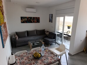 A living room with a sofa, a coffee table, a dining table with chairs, and a sliding glass door leading to an outdoor area.
