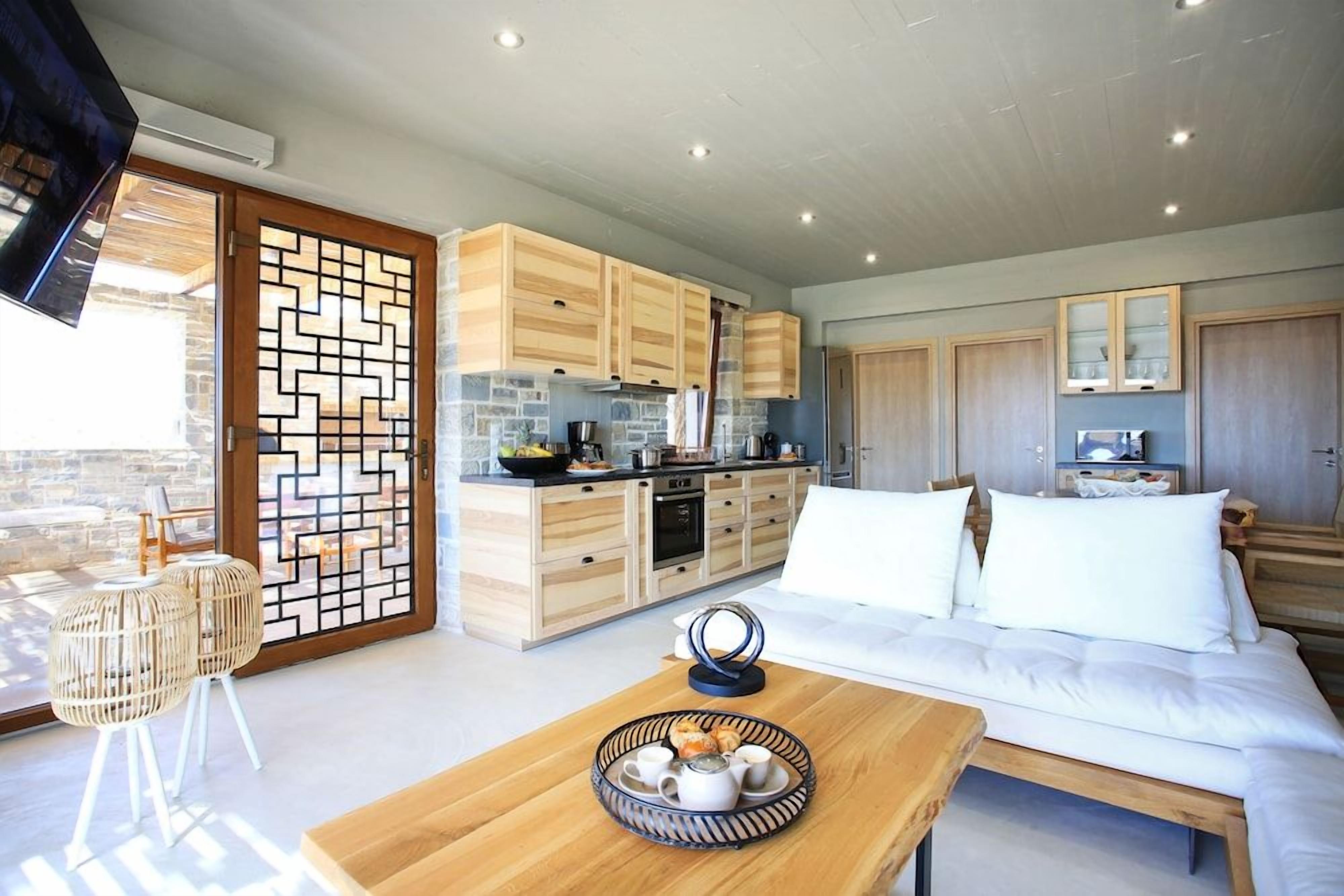 A modern kitchen with wooden cabinets and a stone backsplash, a dining area with a wooden table and chairs, and a bedroom with a large bed and white bedding.