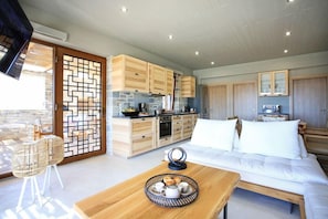 A modern kitchen with wooden cabinets and a stone backsplash, a dining area with a wooden table and chairs, and a bedroom with a large bed and white bedding.