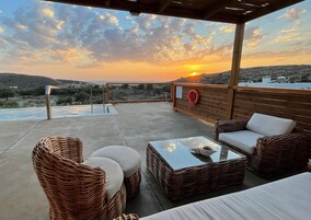 A covered outdoor seating area with wicker furniture, a glass coffee table, and a view of the sunset over a hilly landscape.