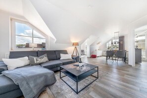 A modern loft apartment with a high ceiling, a living area with a sofa and coffee table, a dining area with chairs, and a kitchen in the background. - Granitz Residential Park 26 Sandschloss (Ostseebad Binz)