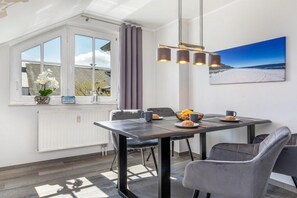 A dining area with a table, chairs, a window with curtains, a radiator, and a painting on the wall. - Granitz Residential Park 26 Sandschloss (Ostseebad Binz)