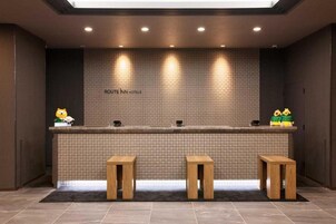 Zona de recepción con pared de azulejos, mostrador de recepción y tres taburetes de madera.
