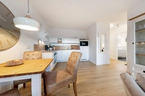 A modern kitchen with white cabinetry, a dining area with a wooden table and leather chairs, and a glass cabinet with wine glasses. - Villa Düne 4 Summer retreat (Ostseebad Binz)