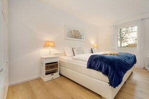 Un dormitorio con cama blanca, dos lámparas de noche, una mesita de noche con espacio de almacenamiento, un cuadro enmarcado en la pared y una ventana con cortinas.