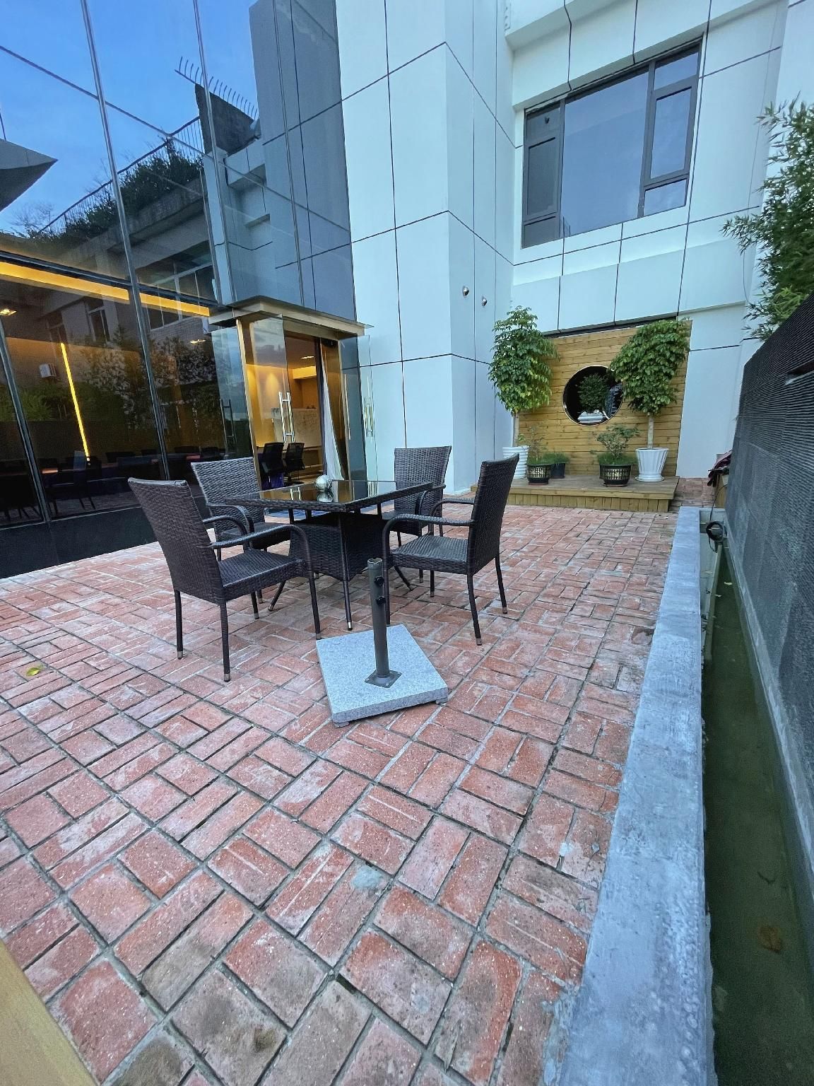 Um pátio externo com mesa e cadeiras, piso de tijolos e parede de vidro.