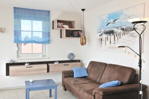 A living room with a brown sofa, a blue table, a window with a blue curtain, a wall-mounted shelf with books, and a painting of birds. - Holiday apartment with 1 bedroom (Graal-Müritz)