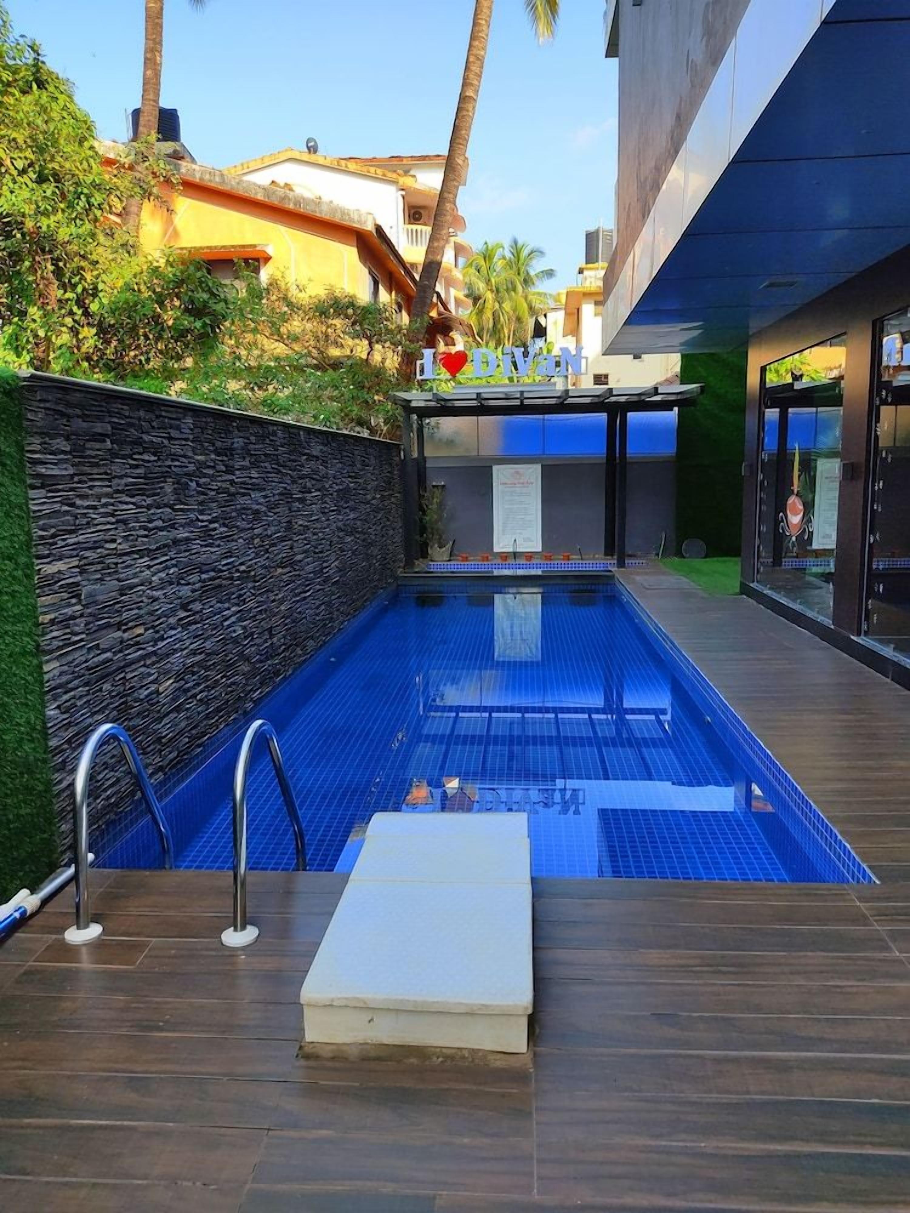 An outdoor swimming pool with a stone wall and wooden deck, surrounded by greenery and buildings.