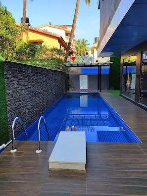 An outdoor swimming pool with a stone wall and wooden deck, surrounded by greenery and buildings.