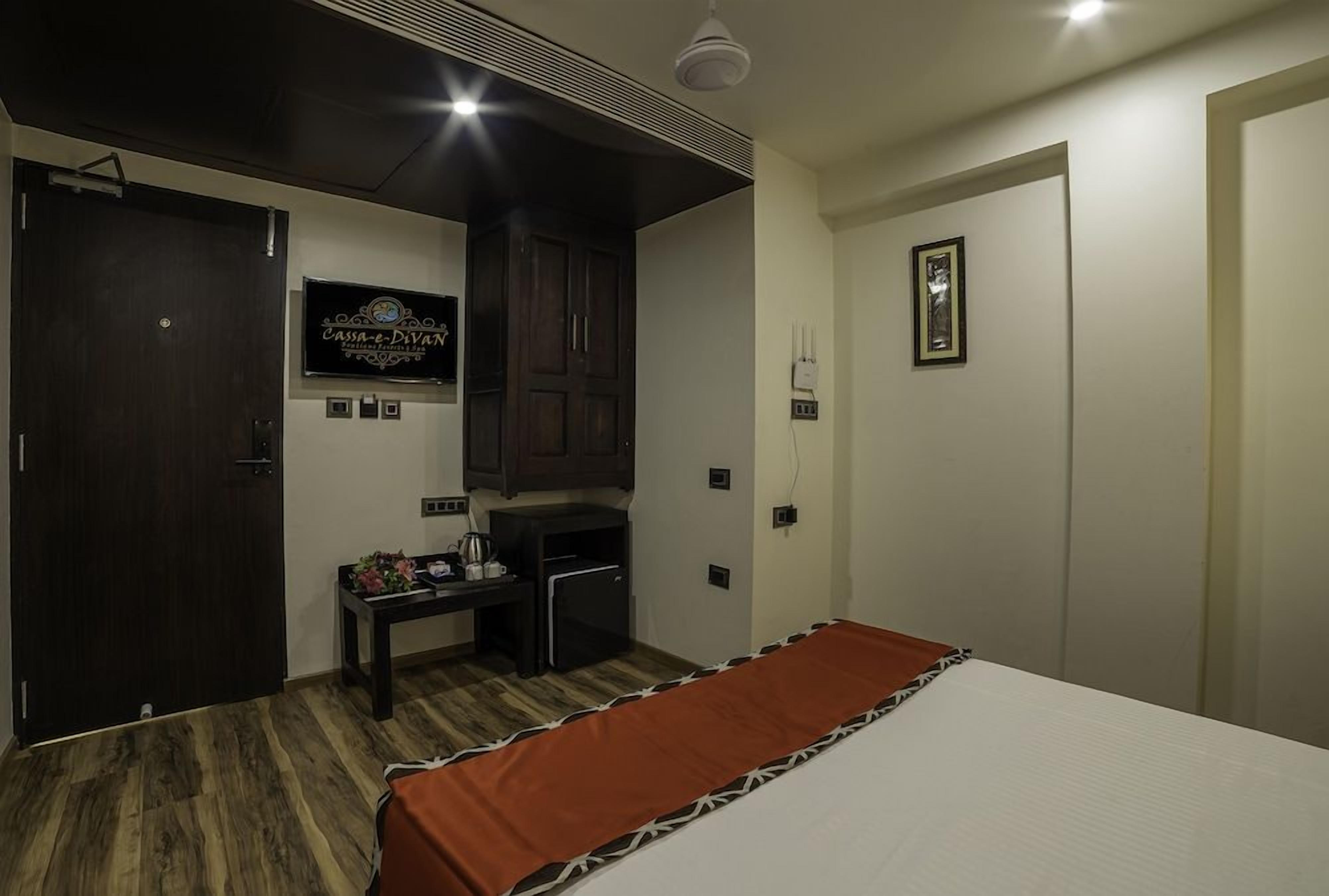 A hotel room with a bed, a small desk with a computer, a dark wooden wardrobe, a television, and a framed picture on the wall.