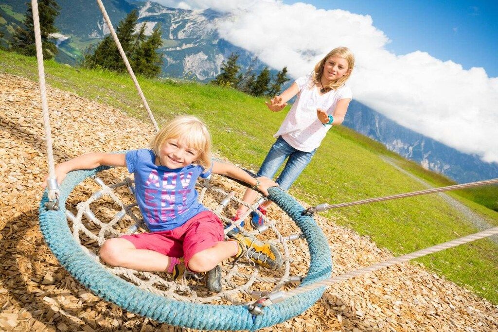 Two children playing on a rope swing in a natural setting with a mountainous backdrop.