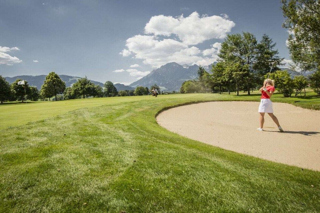 A golf course with a clear sky, trees, and mountains in the background.
