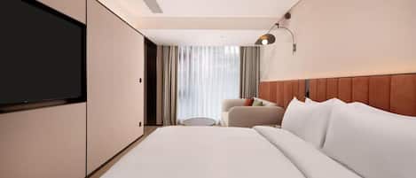 A hotel room with a large bed, a television, a seating area with a chair and a small table, and a window with curtains.