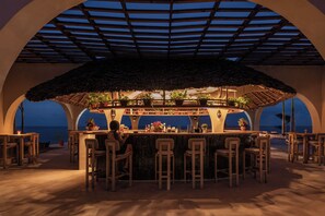 An outdoor bar area with a thatched roof, multiple seating arrangements, and a view of the sea. - Joud by the Sea Zanzibar (Jambiani)