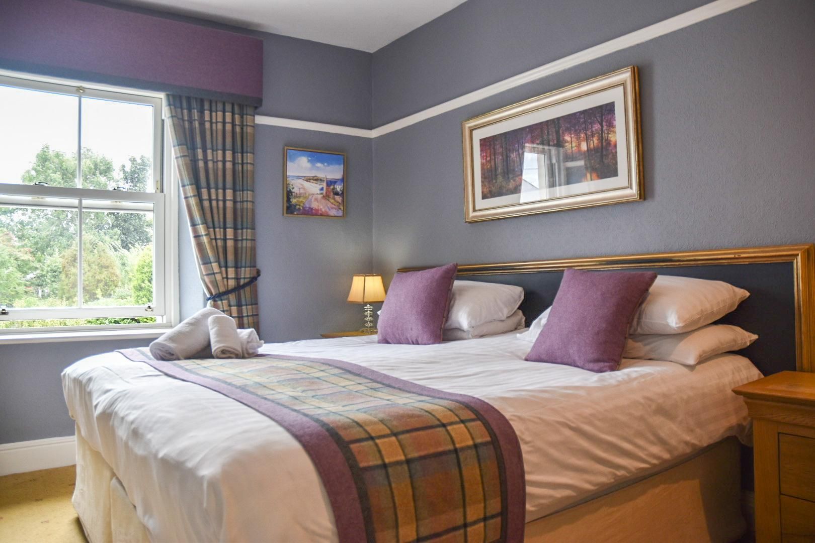 A bedroom with a large bed, a window with curtains, a bedside table with a lamp, and two framed pictures on the wall.
