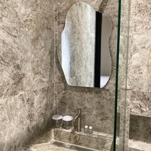 A bathroom with a marble wall, a mirror, a sink, and a glass shower enclosure. - Moonstone (Bandung)