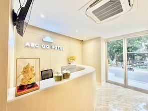Un mostrador de recepción de hotel con un cartel que dice 'ABC QQ HOTEL', un televisor de pantalla plana, una planta en maceta y una obra de arte enmarcada.