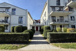 Ein Wohngebäude mit einem zentralen Eingang, mehreren Balkonen und einem gepflegten, von einer Hecke gesäumten Weg.