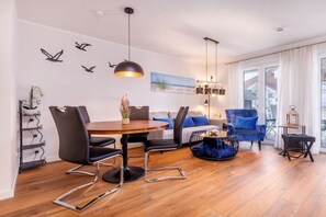 Ein modernes Wohnzimmer mit Essbereich, mit einem runden Holztisch, schwarzen Stühlen, einem blauen Sofa und einer dekorativen Vogel-Wandmalerei.