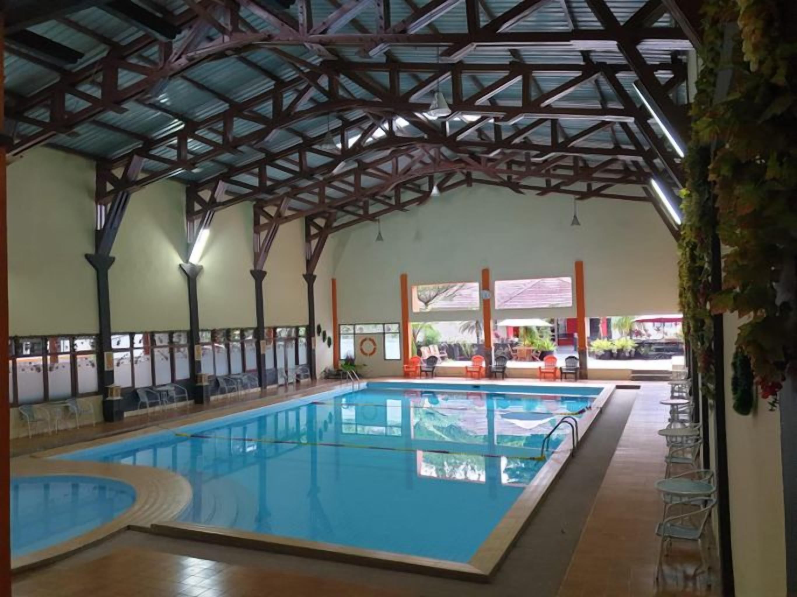 Una piscina interior con techo alto y columnas, rodeada de sillones de descanso y un área de estar cubierta.