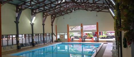 Una piscina interior con techo alto y columnas, rodeada de sillones de descanso y un área de estar cubierta.