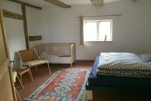 A room with a crib, a chair, a bed with a patterned bedspread, and a window with curtains. - Timber-framed house Melz (Melz)