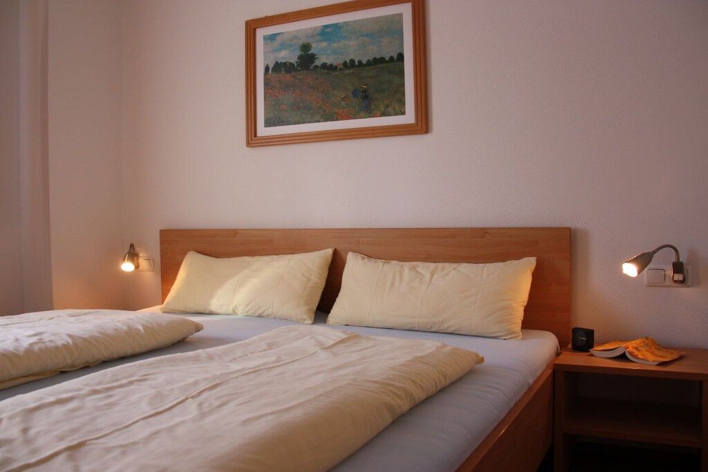 Een tweepersoonsbed met een houten hoofdbord, twee bedlampjes, een ingelijst schilderij aan de muur en een nachtkastje met een lamp en een boek.
