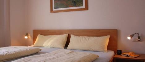 Een tweepersoonsbed met een houten hoofdbord, twee bedlampjes, een ingelijst schilderij aan de muur en een nachtkastje met een lamp en een boek.