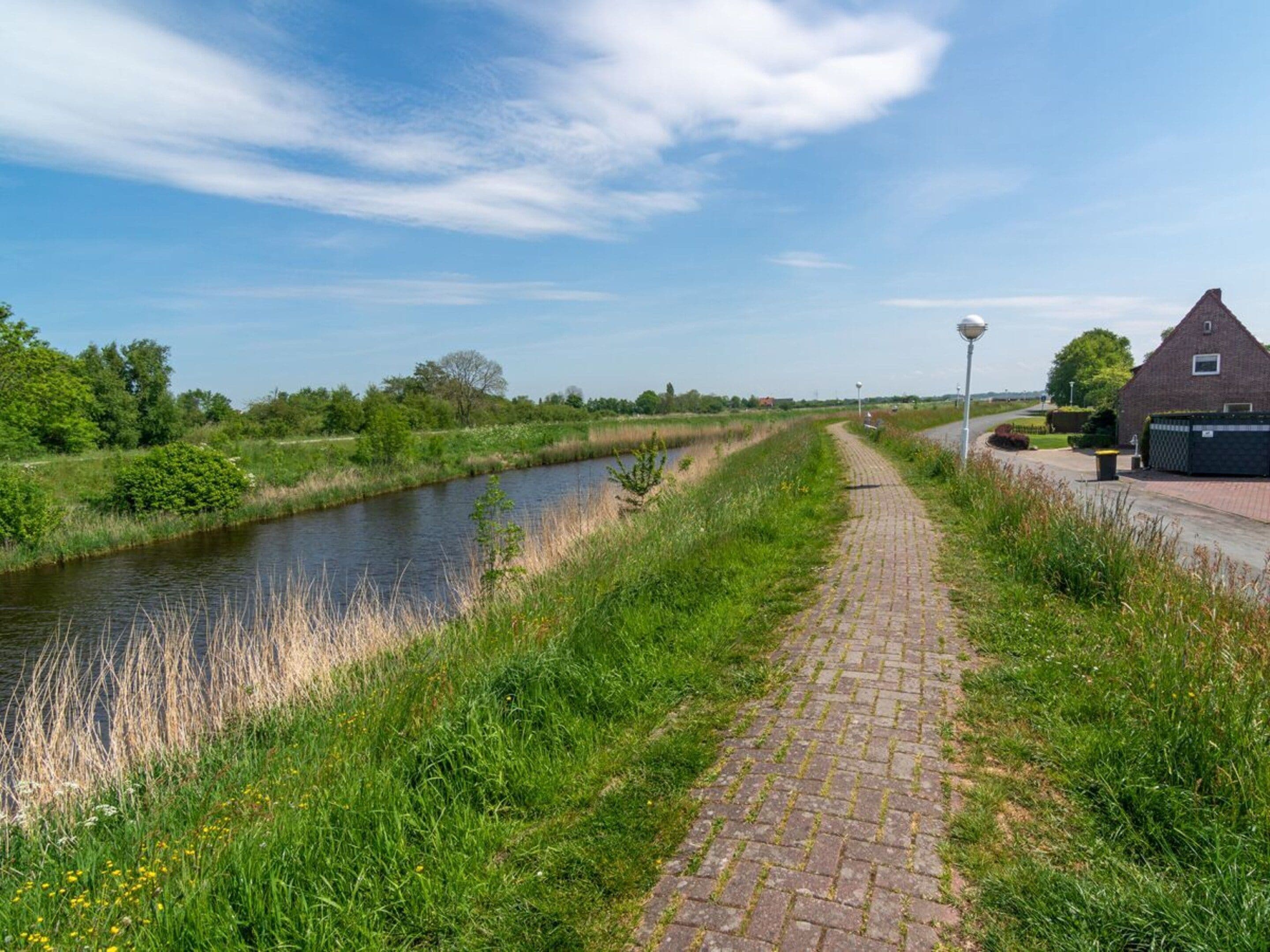 A paved pathway alongside a canal, with a brick building and greenery in the background.