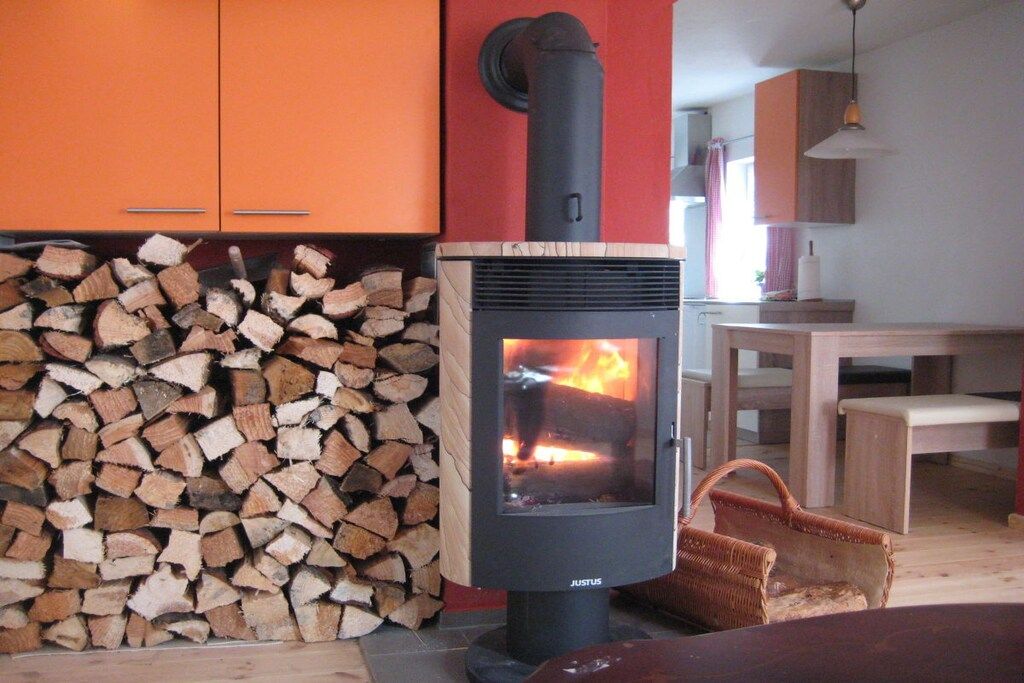 Un poêle à bois avec une flamme visible, une pile de bois de chauffage et un coin repas avec une table et des chaises.