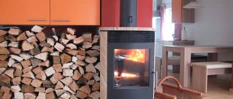Un poêle à bois avec une flamme visible, une pile de bois de chauffage et un coin repas avec une table et des chaises.