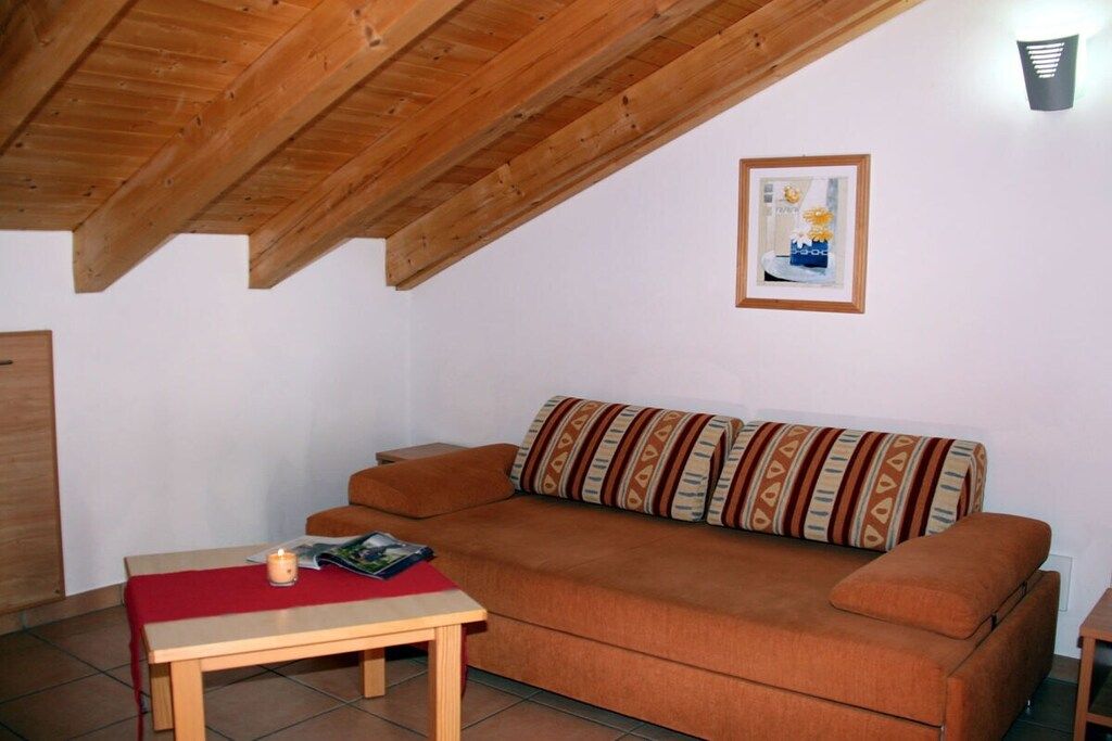 Een gezellige woonkamer met een houten plafond, een bank met opvallende kussens, een bijzettafeltje met een kaars, en een lijst met een afbeelding aan de muur.