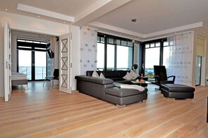 Ein modernes Wohnzimmer mit einer Couch, einem Hocker und einem Couchtisch. Ein großes Fenster bietet einen Blick ins Freie.