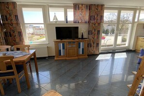Ein Zimmer mit Fernseher, Holzmöbeln, einem Esstisch und großen Fenstern mit Vorhängen.