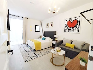 Ein modernes Hotelzimmer mit Bett, Sofa und Couchtisch. Es gibt ein großes Fenster mit Vorhängen, eine Chandelüster und ein Wandbild.