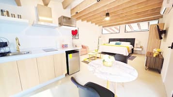 Cozinha moderna com micro-ondas embutido e uma pequena geladeira. Há uma área de refeições com uma mesa redonda e cadeiras, e uma cama com cabeceira ao fundo.