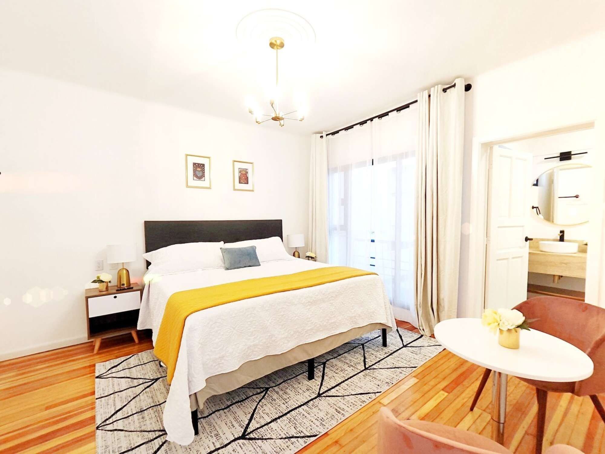 Quarto de hotel com cama, mesa de cabeceira, abajur e uma pequena mesa redonda com uma cadeira.
