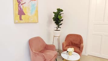 A room with two pink chairs, a small round table with a vase, a potted plant, and a colorful painting on the wall.