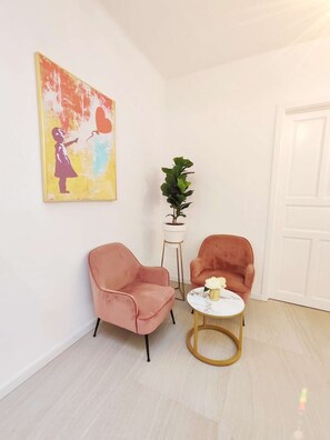 Ein Zimmer mit zwei rosafarbenen Stühlen, einem kleinen runden Tisch mit einer Vase, einer Topfpflanze und einem farbenfrohen Bild an der Wand.