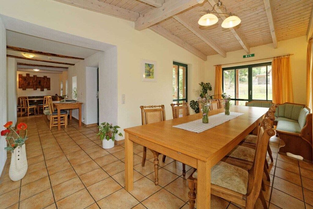 Zona de menjador amb taula i cadires de fusta, cuina al fons i vistes a l'exterior a través d'una finestra gran.