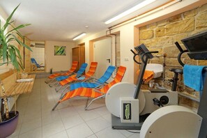 Ein Zimmer mit Trainingsgeräten, darunter ein Ergometer und ein Rudergerät, sowie mehrere farbenfrohe Stühle.