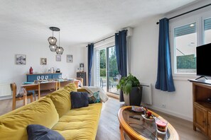 Una sala de estar con un sofá amarillo, una mesa de comedor con sillas, un televisor y un ventanal con cortinas.