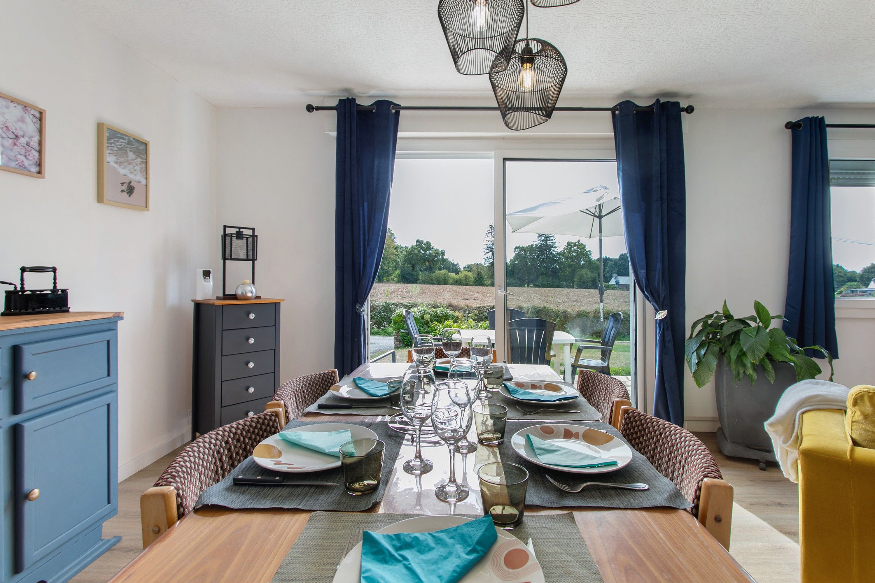 A dining area with a table set for four, a view of the outdoors, and a blue cabinet to the left.