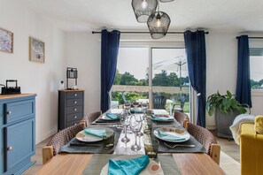 A dining area with a table set for four, a view of the outdoors, and a blue cabinet to the left. - Holiday home 'Petite Maison de Kervenou' with private terrace, garden and Wi-Fi (Goudelin)